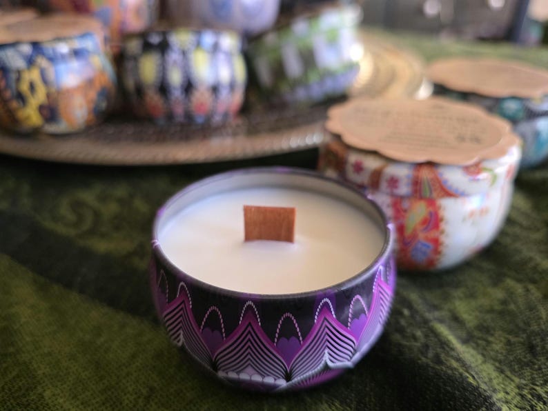 May include: A close-up of a scented candle in a decorative tin. The candle has a wooden wick and is surrounded by a purple and black patterned design. Other candles with colorful designs are in the background, suggesting a collection of candles.