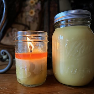 May include: Two mason jars, one with a lit candle and the other filled with a yellow substance. The candle is layered with orange and white wax. The jar on the right has a silver lid and is filled with a creamy yellow substance.