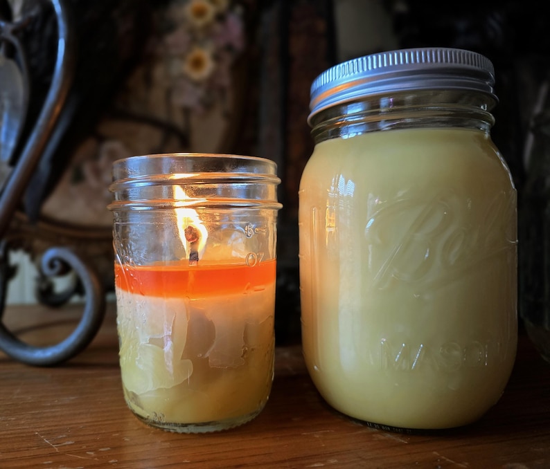 May include: Two mason jars, one with a lit candle and the other filled with a yellow substance. The candle is layered with orange and white wax. The jar on the right has a silver lid and is filled with a creamy yellow substance.