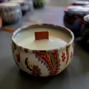 May include: A round candle in a decorative tin. The tin has a cream background with a paisley pattern in red, blue, and green. A wooden wick sits in the center of the white candle wax. Other candles are visible in the background.