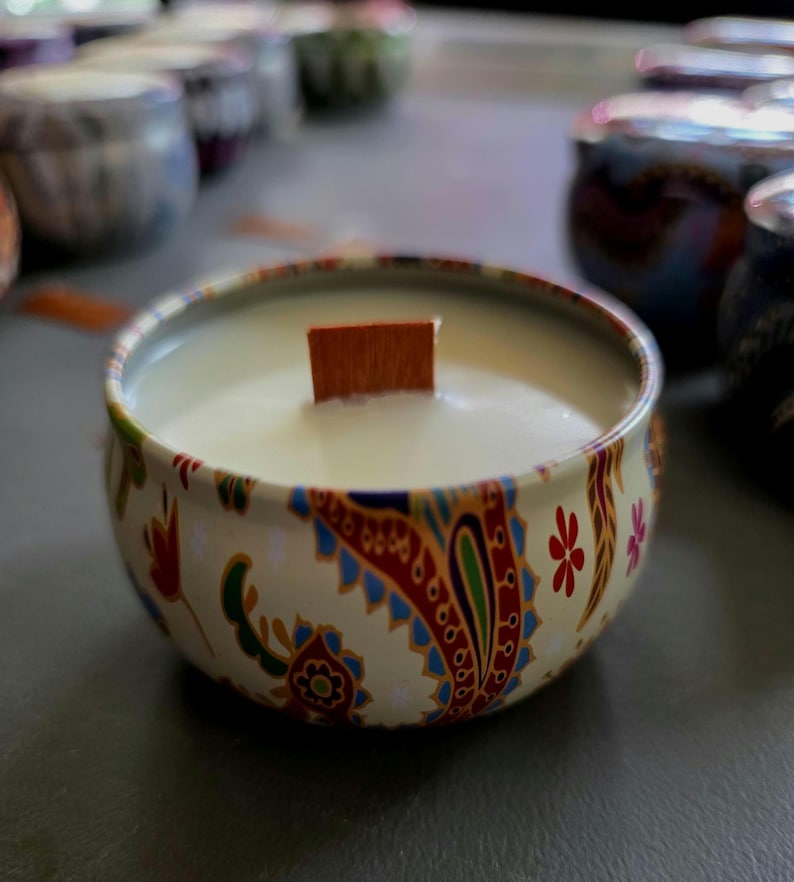 May include: A round candle in a decorative tin. The tin has a cream background with a paisley pattern in red, blue, and green. A wooden wick sits in the center of the white candle wax. Other candles are visible in the background.
