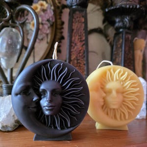 May include: Two round candles with celestial designs. One is black with a sun and moon face, the other is yellow with a similar design. Both candles have a wick at the top and are on a wooden surface.