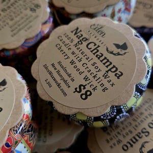 May include: Close-up of several Nag Champa scented candles in decorative tins. Each candle has a paper label with the text "Nag Champa 100% Natural Soy Wax Candle with Crackling Cherry Wood Wick". The tins are 2.5oz.