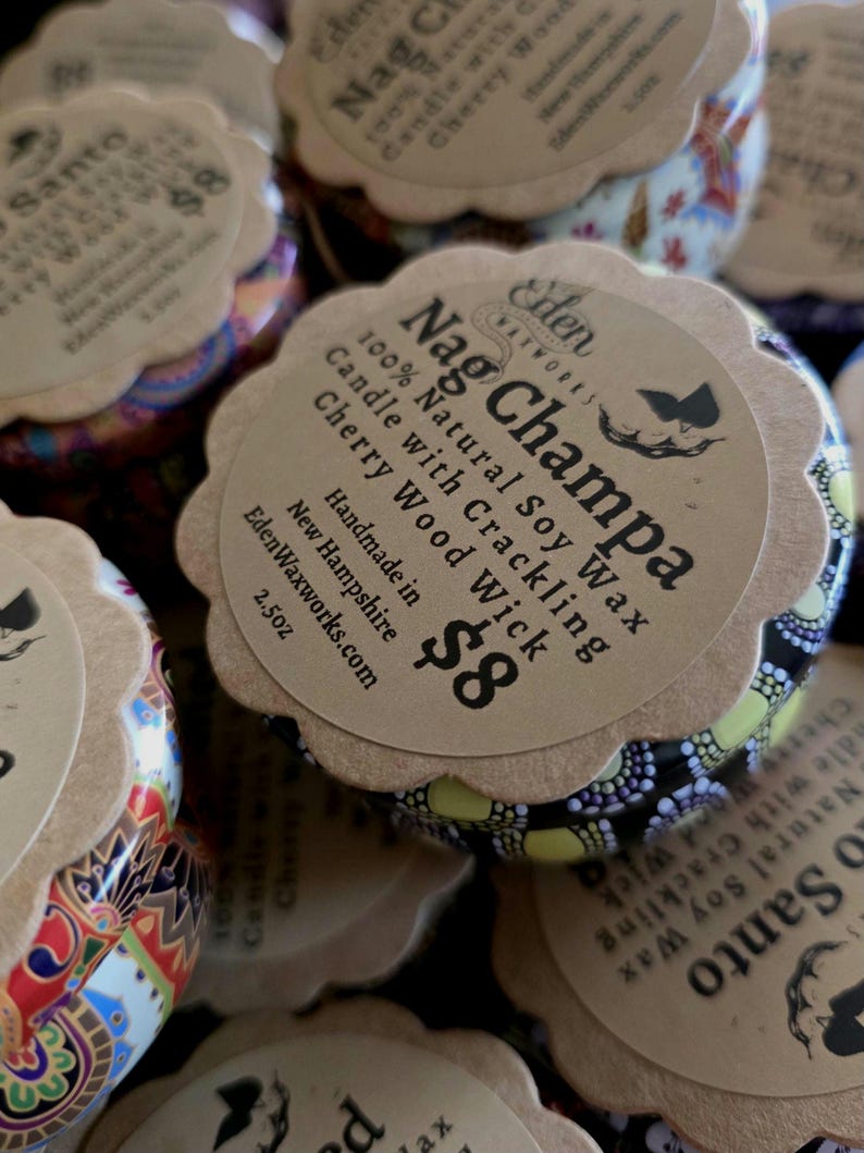 May include: Close-up of several Nag Champa scented candles in decorative tins. Each candle has a paper label with the text "Nag Champa 100% Natural Soy Wax Candle with Crackling Cherry Wood Wick". The tins are 2.5oz.