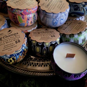 May include: A collection of soy wax candles in decorative tins, each with a wooden wick. The tins feature colorful patterns and labels with scent names like "Nag Champa" and "Palo Santo". The candles are displayed on a silver tray.