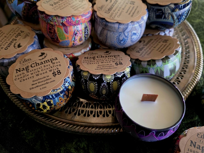 May include: A collection of soy wax candles in decorative tins, each with a wooden wick. The tins feature colorful patterns and labels with scent names like "Nag Champa" and "Palo Santo". The candles are displayed on a silver tray.