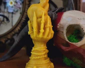 May include: A yellow wax candle shaped like a hand holding a heart. The candle is decorated with intricate details and has a base with a decorative design.