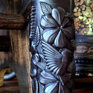 May include: A black pillar candle with a detailed design of a hummingbird and flowers. The candle is decorated with a 3D effect, making the flowers and bird appear to be sculpted.