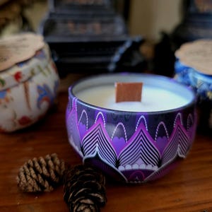 May include: A collection of decorative candles in patterned tins. The central candle is in a purple tin with a geometric design and a wooden wick. Other tins are visible, along with pine cones, on a wooden surface.