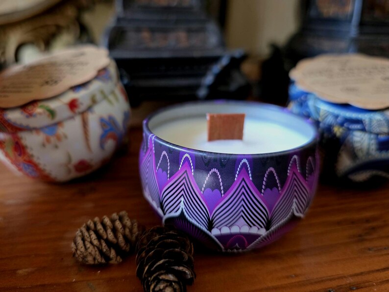 May include: A collection of decorative candles in patterned tins. The central candle is in a purple tin with a geometric design and a wooden wick. Other tins are visible, along with pine cones, on a wooden surface.