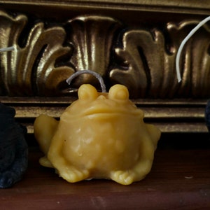 May include: A yellow frog-shaped candle with a wick. The candle has a detailed design, with visible eyes and feet. The candle is made of wax and is sitting on a wooden surface. The background features a gold-colored decorative element.