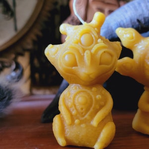 May include: A yellow beeswax candle shaped like a small alien with three eyes and a detailed design. The candle is sitting on a wooden surface.