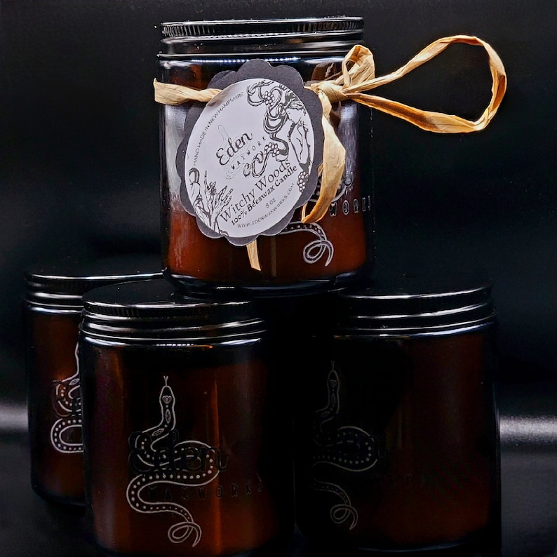 May include: Three brown glass candle jars with black lids. The top jar has a label that reads 'Eden Waxworks' and 'Witchy Woods 100% Soy Wax Candle 8 oz'. The jars have a snake design on the front.