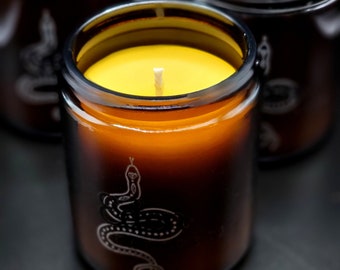 Könnte beinhalten: Nahaufnahme einer Kerze in einem dunkelbraunen Glas. Die Kerze hat gelbes Wachs und einen weißen Docht. Ein weißes Schlangenmuster ist auf das Glas geätzt. Im Hintergrund sind weitere ähnliche Kerzen zu sehen.