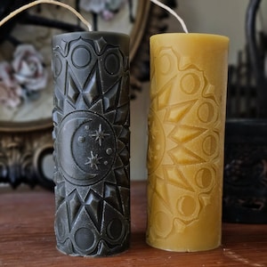 May include: Two tall, cylindrical candles with intricate carvings. One candle is dark green with a moon and stars design, the other is yellow with a sun design.