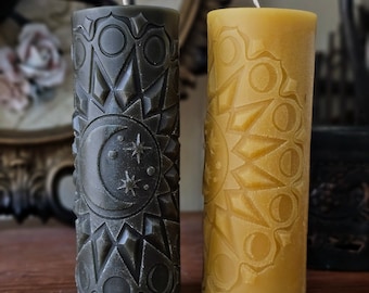 May include: Two tall, cylindrical candles with intricate carvings. One candle is dark green with a moon and stars design, the other is yellow with a sun design.
