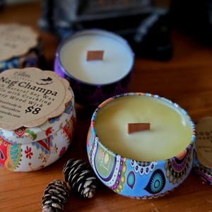 May include: Several decorative candles in patterned tins with wooden wicks are displayed. One candle has a label that reads "Nag Champa 100% Natural Soy Wax Candle with Crackling Cherry Wood Wick". The candles are on a wooden surface.