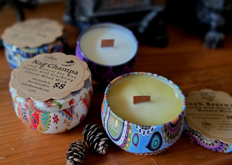May include: Several decorative candles in patterned tins with wooden wicks are displayed. One candle has a label that reads "Nag Champa 100% Natural Soy Wax Candle with Crackling Cherry Wood Wick". The candles are on a wooden surface.