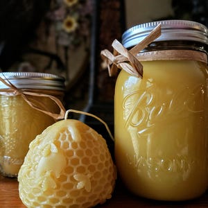 May include: A close-up of three items: two glass jars filled with yellow liquid and a honeycomb-shaped candle. The jars have silver lids and are decorated with raffia. The candle is a light yellow color and has a detailed honeycomb pattern.