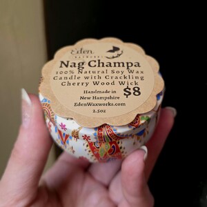 May include: A small, round candle tin with a decorative floral pattern in red, blue, and gold. The label reads "Nag Champa 100% Natural Soy Wax Candle with Crackling Cherry Wood Wick." The candle is handmade in New Hampshire and weighs 2.5 oz.