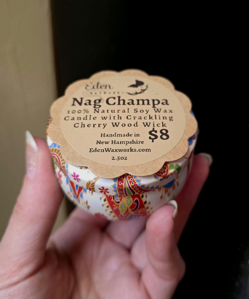 May include: A small, round candle tin with a decorative floral pattern in red, blue, and gold. The label reads "Nag Champa 100% Natural Soy Wax Candle with Crackling Cherry Wood Wick." The candle is handmade in New Hampshire and weighs 2.5 oz.