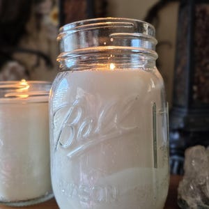 May include: A clear glass Ball Mason jar filled with white wax and a lit candle wick. The embossed text Ball and Mason is visible on the front of the glass container. The candle is illuminated from within, casting a warm glow through the wax.