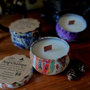 May include: Three decorative candles in patterned tins with wooden wicks. The central candle is in focus, showcasing a colorful paisley design. The label reads "Nag Champa 100% Natural Soy Wax Candle with Crackling Cherry Wood Wick". Two small pinecones are next to the candle.