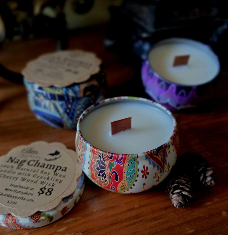 May include: Three decorative candles in patterned tins with wooden wicks. The central candle is in focus, showcasing a colorful paisley design. The label reads "Nag Champa 100% Natural Soy Wax Candle with Crackling Cherry Wood Wick". Two small pinecones are next to the candle.