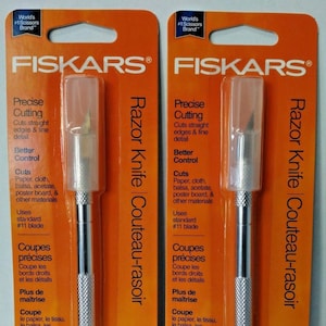 May include: Two orange and white packaging cards for Fiskars Razor Knife Couteau-rasoir. The packaging shows a silver razor knife with a #11 blade. The text on the packaging reads "Precise Cutting", "Better Control", "Cuts", "Uses standard #11 blade", "Coupes précises", "Plus de maîtrise", "Coupe", "Utilise des lames standard n° 11", "Lifetime Warranty | Garantie à vie".