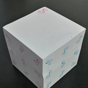 May include: A white cube-shaped notepad with a pink and blue swirl pattern. The notepad has a white top and the sides are covered in a repeating pattern of pink and blue swirls.
