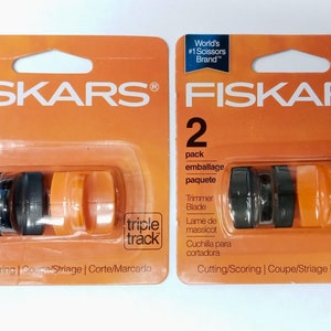 May include: Two packs of Fiskars trimmer blades. Each package is orange and features two black and two orange blades. The packaging includes the text "World's #1 Scissors Brand" and "Trimmer Blade".