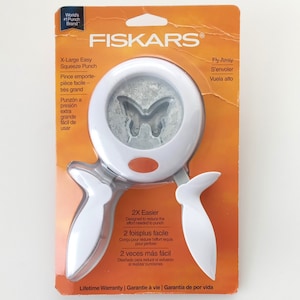 May include: White Fiskars X-Large Easy Squeeze Punch with a butterfly design. The packaging is orange and features the text "Fly Away" in English, French, and Spanish. The punch is designed to reduce the effort needed to punch.