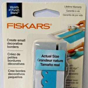 May include: A white and blue decorative border punch cartridge for use with a Fiskars border punch. The cartridge is labeled "Actual Size" and shows a sample of the border design. The cartridge is labeled "Small Cartridge" in English, "Petite Cartouche" in French, and "Cartucho pequeño" in Spanish.