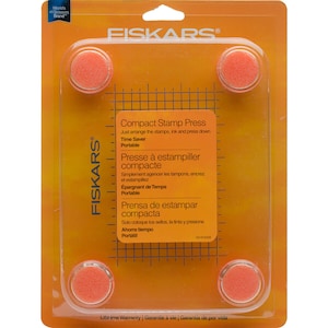 May include: Orange plastic packaging with a clear plastic window showing a Fiskars Compact Stamp Press. The press has four orange foam pads and a gridded surface. The packaging includes text in English, French, and Spanish describing the product's features and benefits.