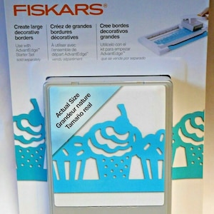 Puede incluir: Un punzón Fiskars blanco y azul con un diseño de cupcake. El punzón está etiquetado como "Tamaño real" y "Cartucho grande".