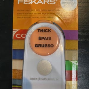 May include: A Fiskars brand paper punch for thick materials, in a clear plastic package. The punch has a white body with an orange circular accent that reads "THICK, ÉPAIS, GRUESO". The packaging is orange and includes text in English, French, and Spanish.