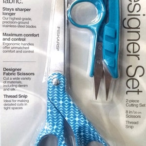 May include: A set of two Fiskars fabric scissors. The larger scissors are 8 inches long and have a teal and white geometric pattern on the handles. The smaller scissors are 2 inches long and have a teal handle. The packaging says "Designer Set" and "2-piece Cutting Set".
