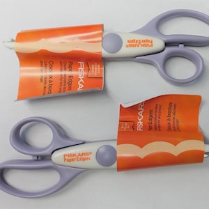 May include: Two pairs of purple and white Fiskars Paper Edgers scissors with orange packaging. The packaging has the text "Fiskars Paper Edgers" and "Ciseaux à bord" in English and French.
