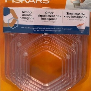 May include: A set of 12 clear plastic hexagon-shaped templates for creating various sizes of hexagons. The templates are nested inside each other and are packaged on a bright orange background with the Fiskars logo and the text "Simply create hexagons".