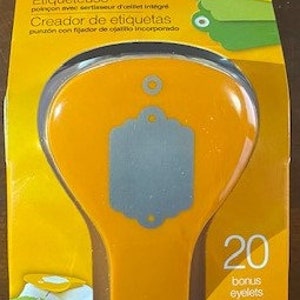 May include: Orange Fiskars tag maker with a built-in eyelet setter. The tag maker is shaped like a light bulb and has a gray tag shape in the center. The packaging includes 20 bonus eyelets. The text on the packaging reads "Tag maker punch with built-in eyelet setter. Etiqueteuse poinçon avec sertisseur d'oeillets intégré. Creador de etiquetas punzón con fijador de ojalillos incorporado. 20 bonus eyelets. lames de ceillets. ojalillos de bonificación."