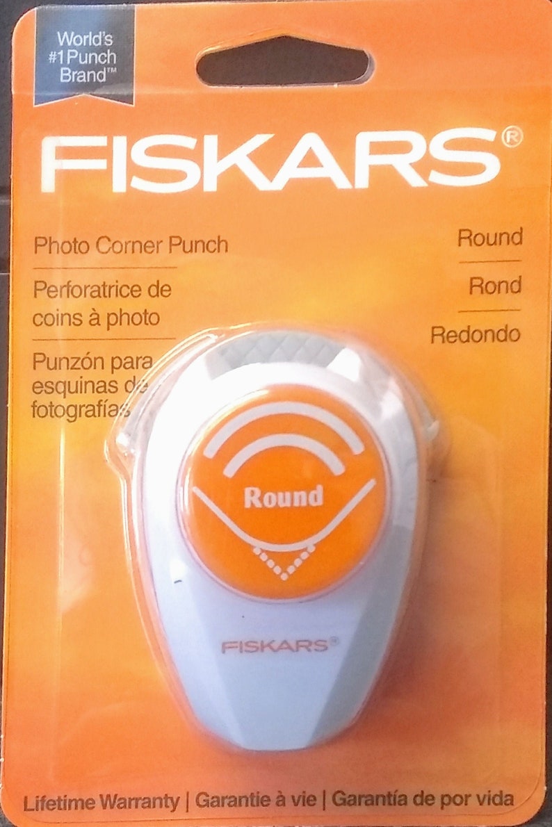 May include: A white plastic photo corner punch with an orange button that says "Round". The packaging is orange and says "FISKARS&reg;" in white letters. The packaging also says "Photo Corner Punch" and includes the words "Round", "Rond", and "Redondo" in different languages.