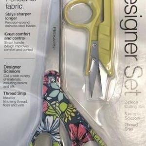 May include: A Fiskars Designer Set containing one pair of 8-inch scissors with a multi-colored floral pattern on the handles and one yellow thread snip. The items are packaged in a clear plastic blister pack on a white cardboard backing.