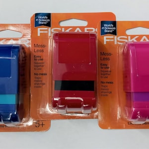 May include: Three packages of Fiskars brand children's scissors. The scissors are in blue, red, and pink. The packaging says "Mess-Less" and "Easy to use".