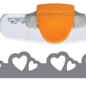 May include: A Fiskars heart-shaped paper punch with an orange handle and the brand name printed on it. The punch is white and gray, with a grid pattern. A gray paper strip with heart cutouts is below the punch.