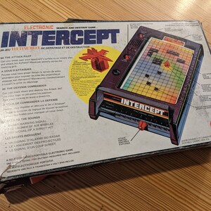 以下が含まれることがあります： レイクサイド製の電子ゲーム「インターセプト」のビンテージボックス。ボックスは青、赤、黄色のカラーで、ゲームボードの画像と英語とフランス語の指示が書かれています。このゲームは、プレイヤーがアタックジェットを追跡して破壊する必要がある、捜索と破壊のゲームです。