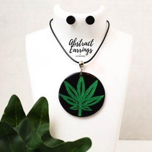 Puede incluir: Un collar negro con un colgante de hoja de marihuana verde y pendientes de botón negros a juego. El collar y los pendientes están en un expositor blanco. El texto "Abstract Earrings & Accessories" está impreso en el expositor.
