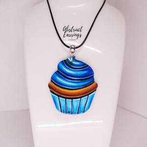 Pode incluir: Um colar com um pingente de cupcake azul e laranja com um cordão preto. O pingente é feito de resina e tem um acabamento brilhante. O colar é um acessório divertido e caprichoso.
