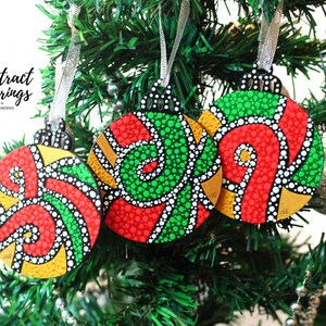 Könnte beinhalten: Drei farbenfrohe, abstrakte Weihnachtskugeln mit roten, grünen, gelben und weißen Punktmustern hängen an einem grünen Weihnachtsbaum. Die Kugeln haben silberne Bänder zum Aufhängen. Der Text "Abstract Earrings" ist sichtbar.