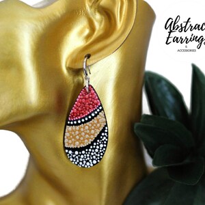 May include: A pair of teardrop-shaped earrings with a black, white, gold, and red abstract design. The earrings are made of painted wood and have silver hooks.