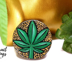 May include: A circular earring featuring a green cannabis leaf design on a black background, encircled by gold dots. The image includes the text "Abstract Earrings & Accessories". The background has gold and green elements.